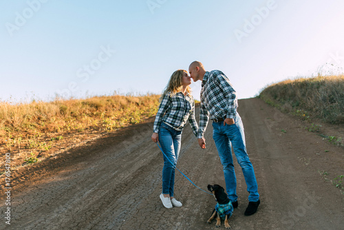 man guy in checkered shirt and girl in green dressed in a black shirt and jeans kiss standing in grass sitting in grass half on top of sunset with dog running around fooling around
