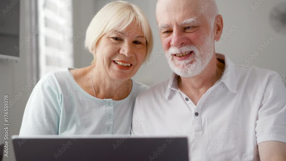 Senior couple sitting at home talking via messenger app Skype. Smiling waving hands in greeting. Remote communication with children on retirement, active modern lifestyle Senior couple sitting at home talking via messenger app Skype. Smiling waving hands in greeting. Remote communication with children on retirement, active modern lifestyle