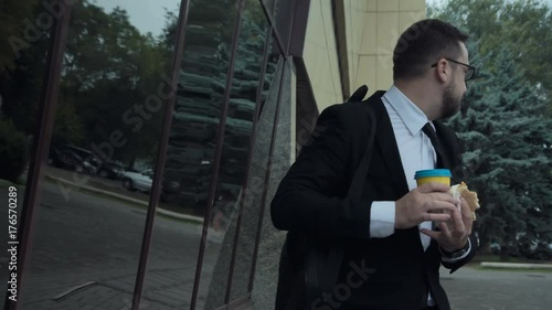 Young angry busy man with an unhealthy lifestyle eating a sandwich, drinking coffee and talking on mobile phone while walking in a hurry on the street