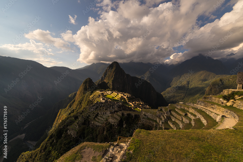 Naklejka premium Machu Picchu illuminated by the warm sunset light. Wide angle view from the terraces above with scenic sky and sun burst. Dreamlike travel destination, world wonder. Cusco Region, Peru.