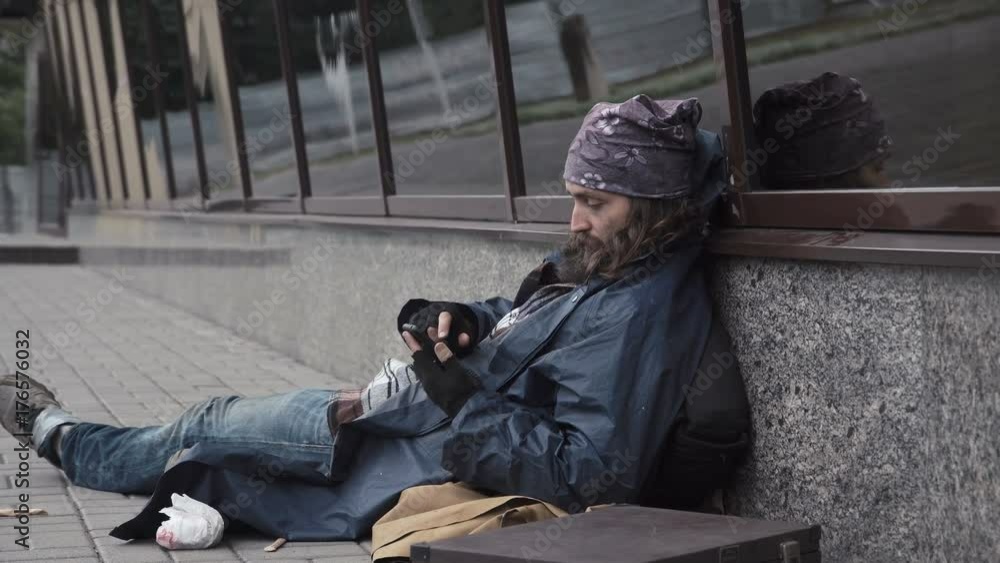 Cheerful homeless man sitting on pavement and using smartphone on street. Cheerful homeless man sitting on pavement and using smartphone on street.