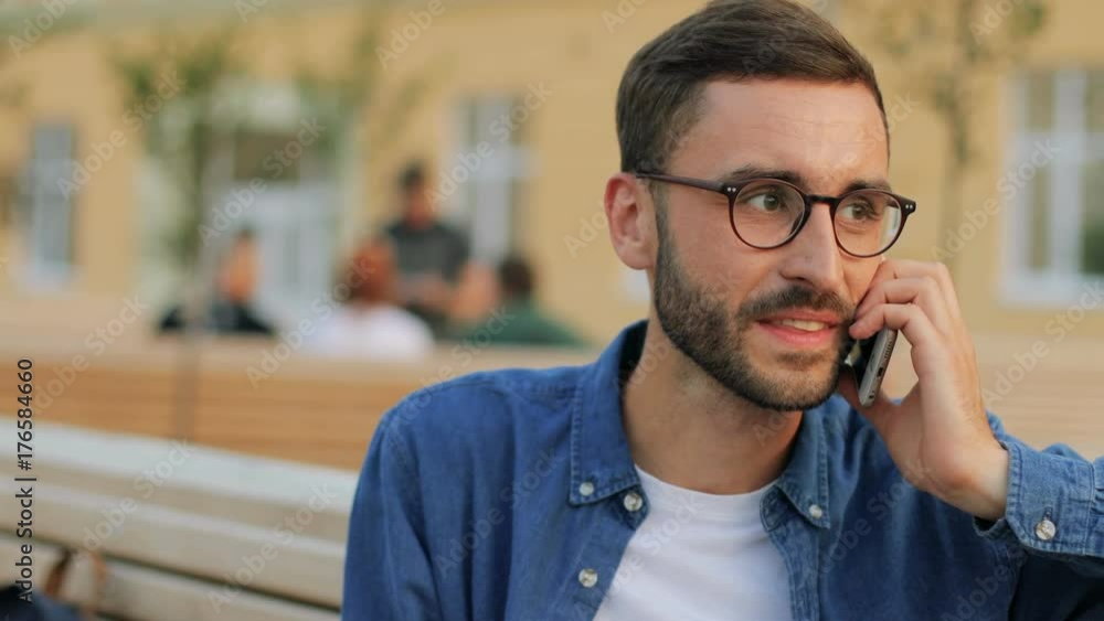 A portrait of an attractive young man talking on the phone - smiling and laughing. Outdoors in the park. Blurred background