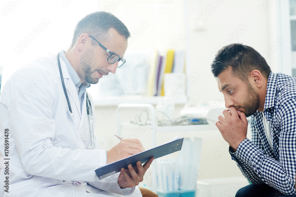 Pensive patient sitting in front of his doctor filling in medical form