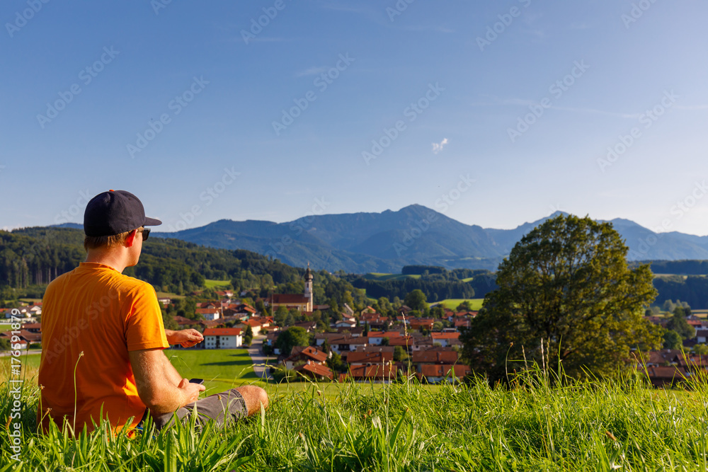 Young Man sitting in meadow overwatching bavarian town with smartphone in hands, with alps in background, low focus