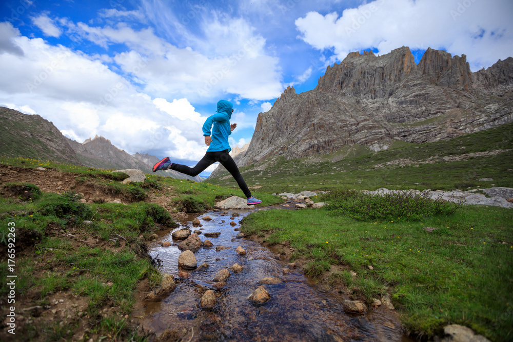 Obraz premium young woman trail runner jumping over stream water at mountain