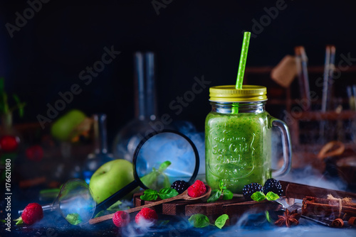 Smoothie jar with green helthy drink in a still life with berries, cinnamon, anise, magnifying glass and nutrition laboratory equipment. Science concept. Dark food photography.