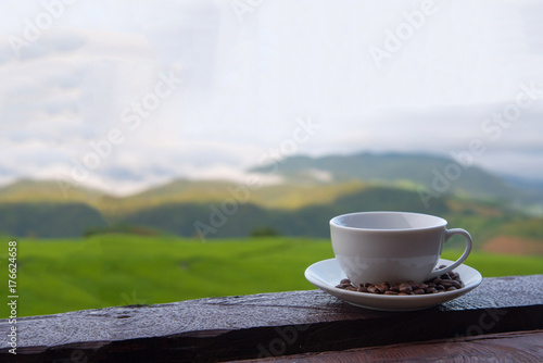 A white cup of hot coffee and coffee beans put interspersed in dish on wood table with green rice field background,wallpaper 