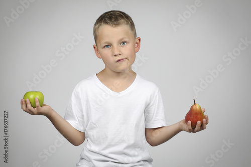 kleiner, blonder Junge hält zweifelnd einen Apfel und eine Birne in der Hand, vor neutralem Hintergrund