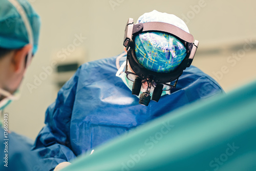 Surgeon doctor operating using special lamp lighting and glasses loupes wearing blue surgical mask and surgical cap in surgery room with his team  surgeons operating photo