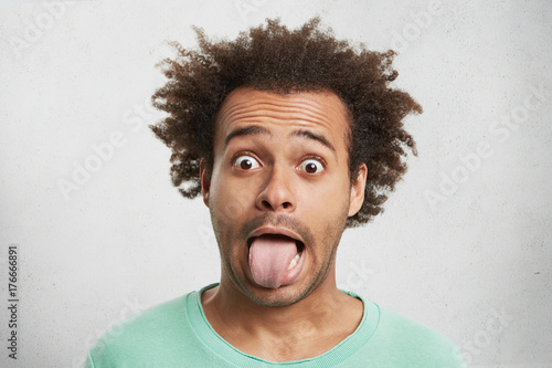 Young funny Afro American man looks surprisely, makes grimace, shows tongue as quarrels with wife. Silly mixed race man with playful expression stares at camera, foolishes alone. Facial expressions photo