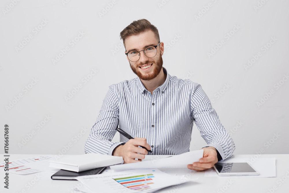 Busy smiling bearded male enterpreneur holds pen and document, has pleased expression as signs successful contract with partners, sits at work place. Glad positive young freelancer works alone Busy smiling bearded male enterpreneur holds pen and document, has pleased expression as signs successful contract with partners, sits at work place. Glad positive young freelancer works alone