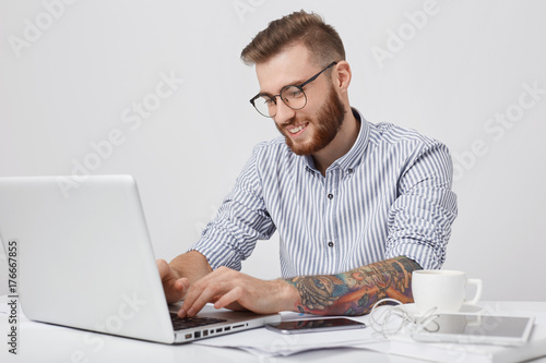 Glad fashionable man with smile, types on generic laptop, checks e mail or messages online, uses high speed wireless internet connection. Successful young male freelancer works remotely at office photo