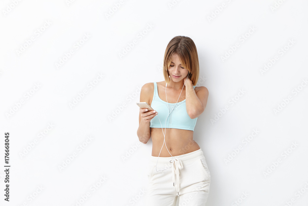 Fitness and technology concept. Restful female model wears sport clothes, listens to favourite music on earphones and smart phone, isolated over white background with copy space for your text Fitness and technology concept. Restful female model wears sport clothes, listens to favourite music on earphones and smart phone, isolated over white background with copy space for your text