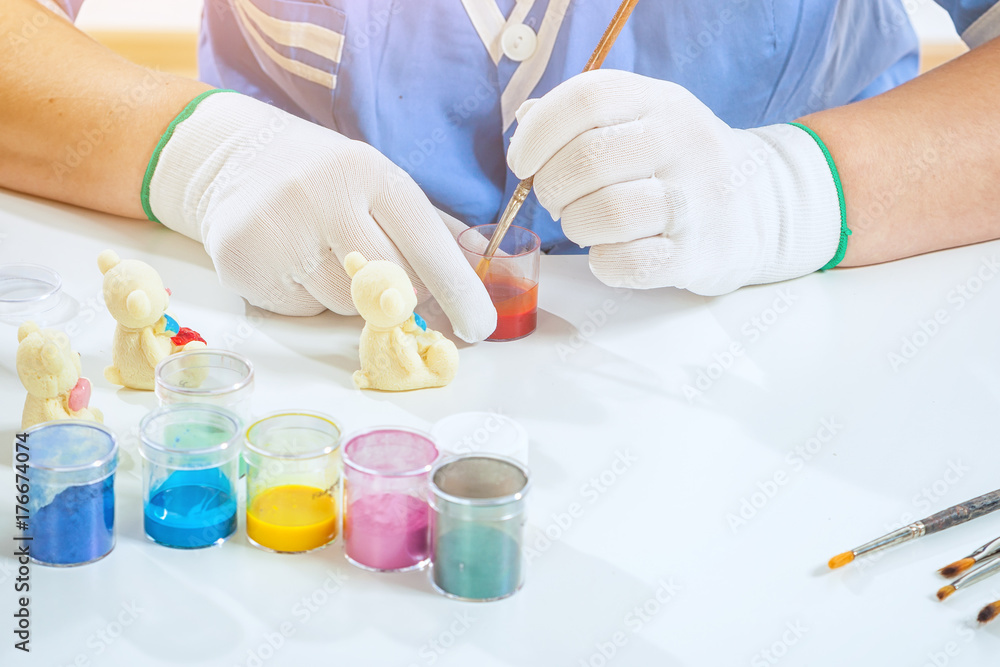 A female confectioner in blue uniform paints a wooden brush with an edible red paint of a white chocolate bear on a white table, on which there is a set of paints and brushes A female confectioner in blue uniform paints a wooden brush with an edible red paint of a white chocolate bear on a white table, on which there is a set of paints and brushes