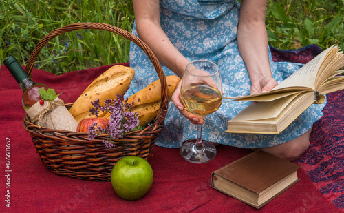 Young woman in romantic blue dress at a picnic. Girl is reading a book and drinking white wine at nature