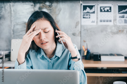 Stressed Asian creative designer woman cover her face with hand and feel upset while talk on mobile phone with customer in front of laptop computer on desk at office,Stress office lifestyle concept