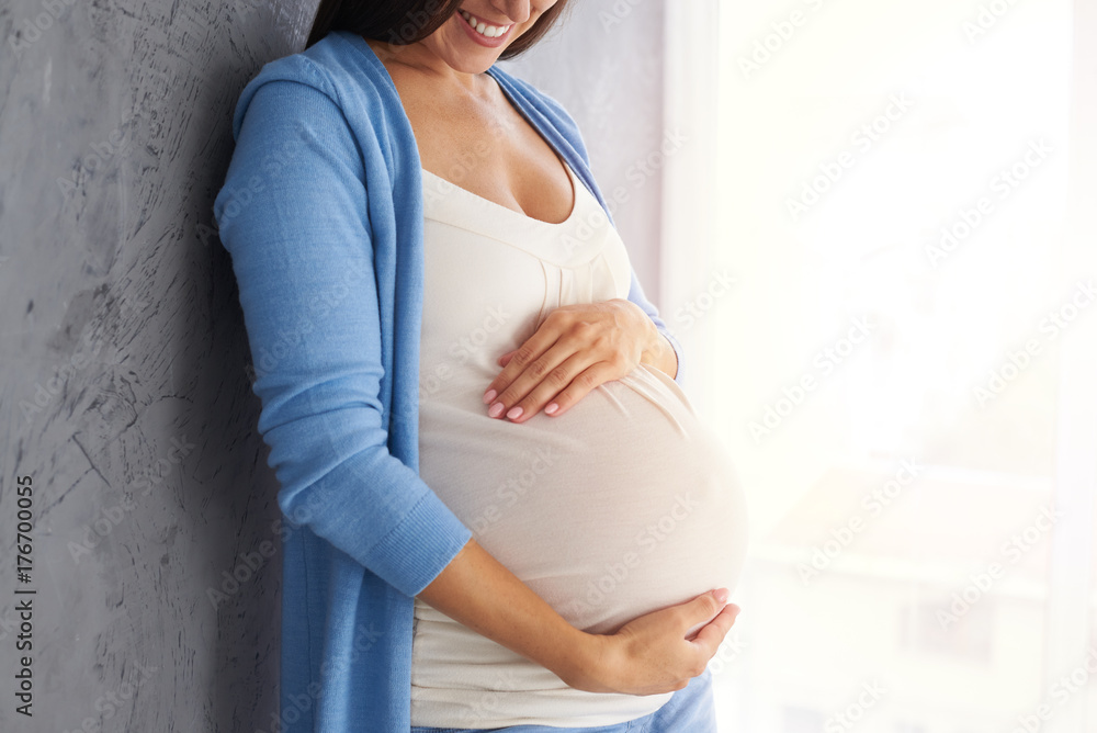 Smiling pregnant woman holding belly while posing against grey