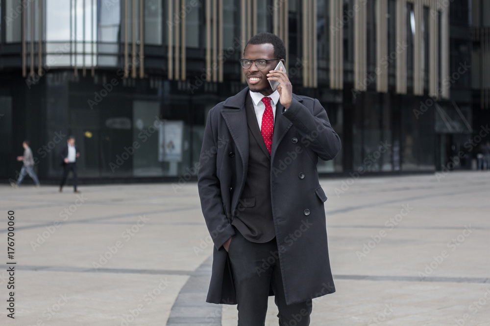 Outdoor image of young African American man moving from one office to another, standing in street in city center, having phone chat about crucial business matters, trying to find solution to problems