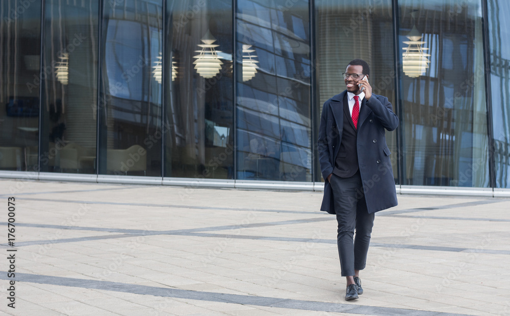 Full length image of African American guy walking outdoors in city center dressed in formal suit and coat, holding smartphone and having pleasant conversation about business or family issues, smiling Full length image of African American guy walking outdoors in city center dressed in formal suit and coat, holding smartphone and having pleasant conversation about business or family issues, smiling