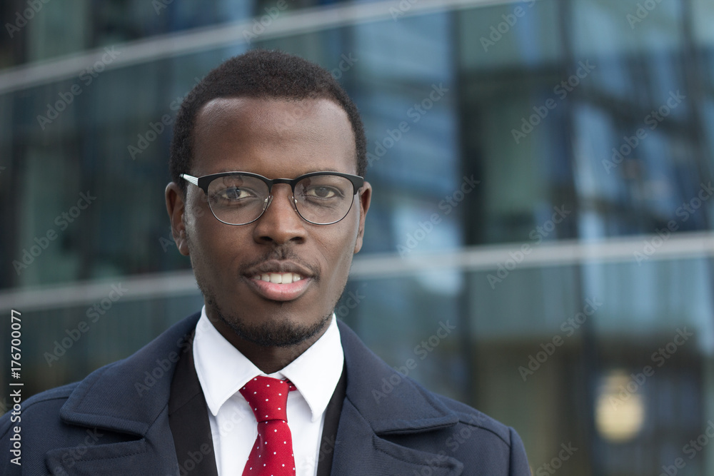 Outdoor closeup of African American entrepreneur pictured in street with high glass building in background, wearing black-rimmed eyeglasses, looking calm but concentrated on business matters