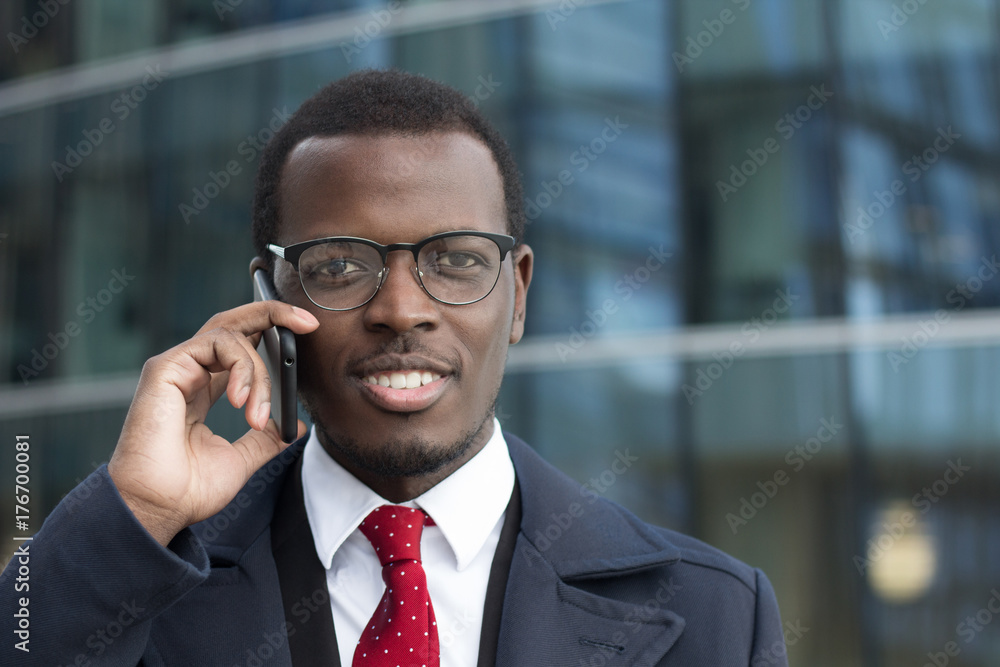 Horizontal portrait of young dark-skinned business guy dressed in formal clothes while standing in street in city center having discussion on cellphone, looking attentive and focused on news he hears Horizontal portrait of young dark-skinned business guy dressed in formal clothes while standing in street in city center having discussion on cellphone, looking attentive and focused on news he hears