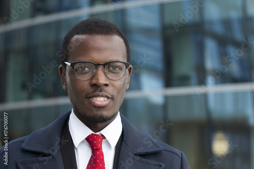 Outdoor closeup of African American entrepreneur pictured in street with high glass building in background, wearing black-rimmed eyeglasses, looking calm but concentrated on business matters