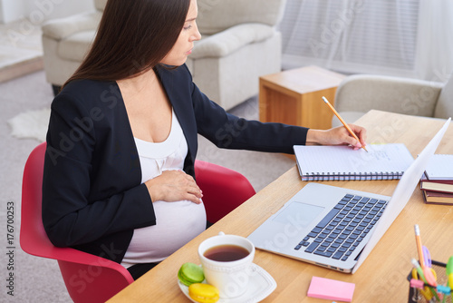 Serious pregnant businesswoman making notes while working on laptop