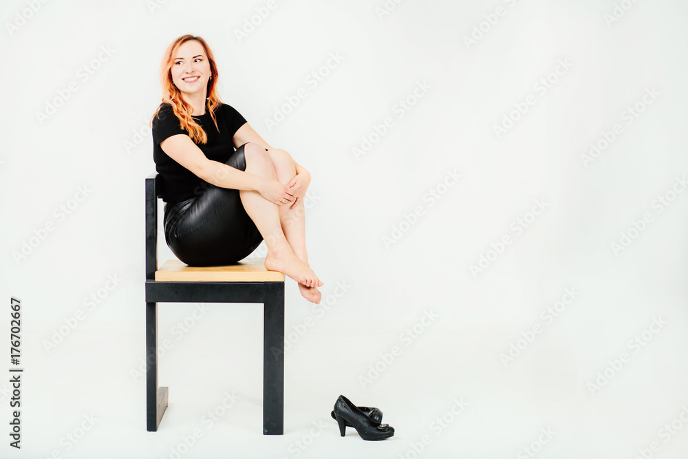 Happy smilling girl in black t-shirt and leather skirt sitting on stool tuck  legs up on white background in studio. Black shoes to lie near her Stock  Photo