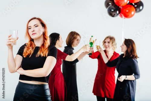 Sad faced woman with glass of champagne drinking alone while her four girl friends with red and black balloons celebrate hen-party.