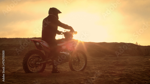 Professional FMX Motorcycle Rider Rests on His Bike and Overlooks Hard Sandy Off-Road Terrain. Sun is Setting.