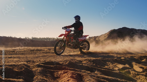Side View Footage of the Professional Motocross Motorcycle Rider Driving on the Dune and Further Down the Off-Road Track. It's Sunset and Track is Covered with Smoke/ Dust/ Dirt.