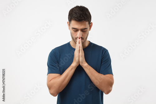 Picture of young guy dressed casually isolated on grey background, having put hands together in prayer or meditation, looking relaxed and calm, dreaming and waiting for all best