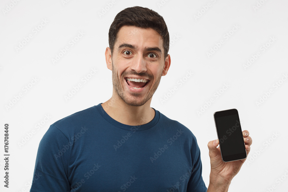 Indoor portrait of young male isolated on gray background presenting blank cellphone and smiling with open mouth with astonishment. Copyspace for advertising goods and services. Indoor portrait of young male isolated on gray background presenting blank cellphone and smiling with open mouth with astonishment. Copyspace for advertising goods and services.