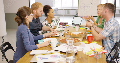 Young multiracial group of people posing at wooden table in office and eating ordered pizza while enjoying conversation.