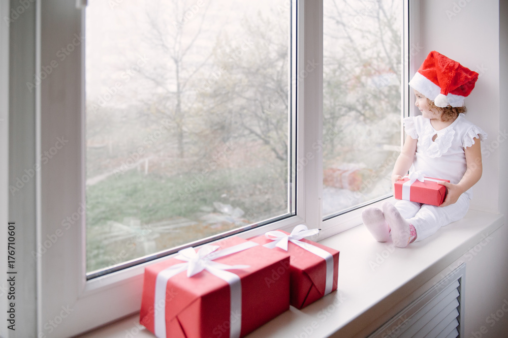 Fototapeta premium little girl in Santa hat sits on window sill and holding gift