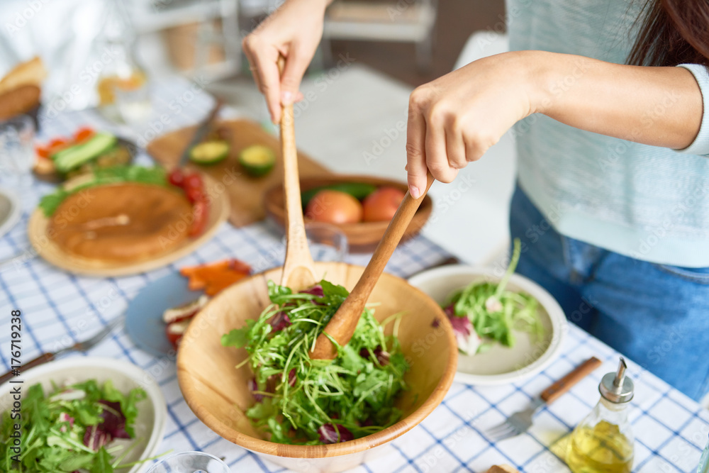 Closeup of young woman mixing green salad in wooden bowl at table with wholesome food while preparing family dinner