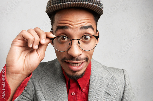 Surprised young dark skinned male with beard and mustache, keeps hand on frame, wears old fashionable cap and jacket, being astonished to hear intriguing news, isolated over white background
