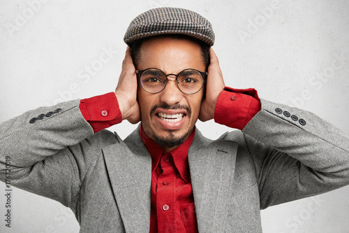 Stressful bearded man in cap and jacket, covers ears, protects himself from noise, doesn`t want to hear too loud sound. Annoyed nerd male being on noisy party, listens music. Negative human expression