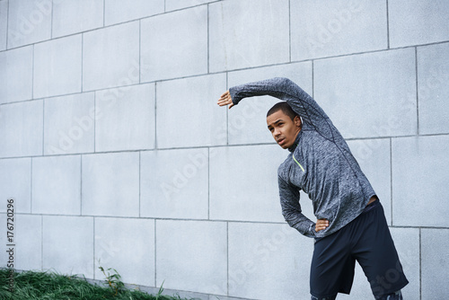 Isolated outdoor portrait of confident young dark-skinned sportsman in stylish outfit bending sideways, doing stretching and warm up exercises, preparing his mucles for workout. Sports and motivation photo