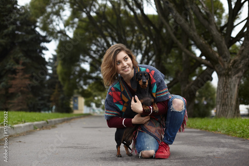 People, pets, leisure and activity. Cheerful young female in ripped jeans and warm plaid sitting on pavement on one knee, embracing her faithful four legged friend. Owner and pet dog resting outdoors
