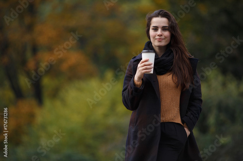 Young stylish woman wearing knitted sweater and coat walking in the autumn park and drinking take away coffee in paper cup