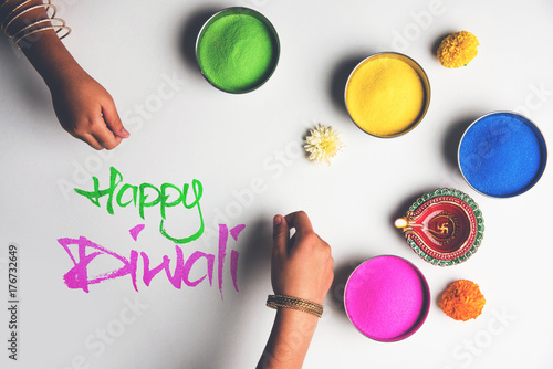 Stock Photo of happy diwali greeting card clicked using elements of Diwali festival like colourful rangoli in bowls, diwali clay lamp or diya and girl or girl making rangoli, writing happy diwali

