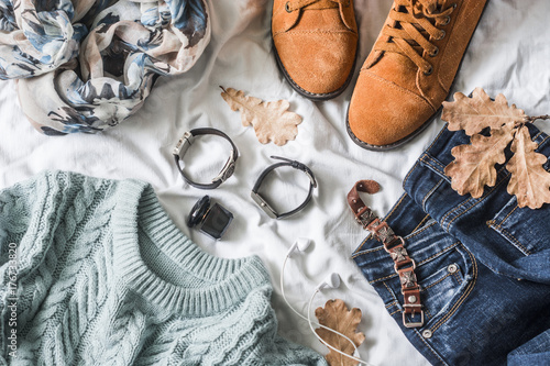 Flat lay women's clothing for autumn walks, top view. Brown suede boots, jeans, a blue pullover, scarf, bracelets, watches, headphones, perfume on a light background. Fashion concept