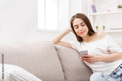 Young brunette woman using smartphone at home on the couch. Dark-haired girl in casual texting online
