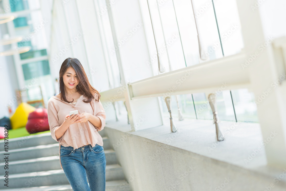 Happy beautiful Asian woman using smartphone while walking in modern office building Happy beautiful Asian woman using smartphone while walking in modern office building