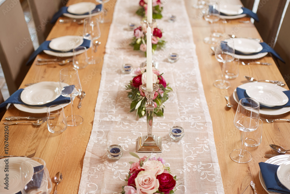 Tischdekoration mit kleinen Blumen, Kerzen und einem weisen Tischläufer auf  einem Holztisch in einem großen offenem Raum. Gedeckt sind weise Teller auf  blauen Servietten mit silbernen Geschirr. Stock Photo | Adobe Stock