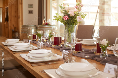 Tischdekoration mit Blumenstrauß und roten Kerzen auf einem grauen Tischläufer auf einem Holztisch in einem großen offenem Raum. Gedeckt ist klassisches weises Geschirr.