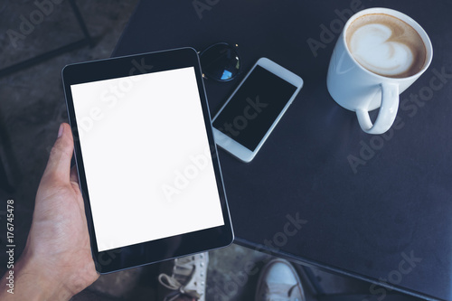 Top view mockup image of a man's hands holding black tablet pc with blank white screen ,coffee cup , mobile phone and sunglasses on a table in cafe background