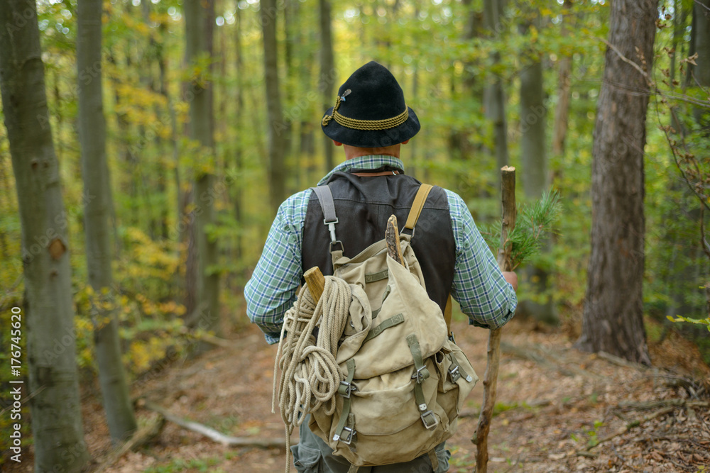An old man in a hat with a backpack
on holiday in the mountains. The elder is leaning against a stick in the autumn forest.

