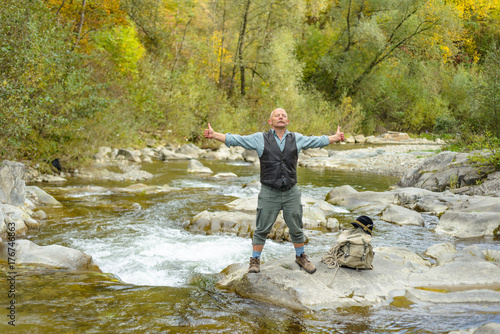 An old man enjoys nature in the outdoors in the mountains. The elder man on the bank of the river raised his hands up. 